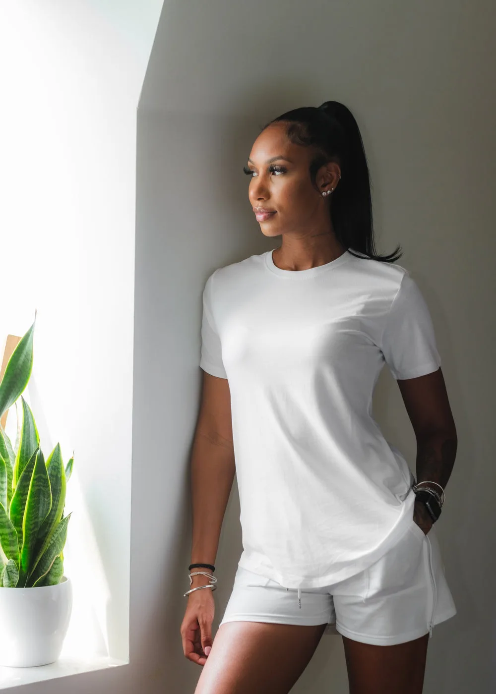 A woman wearing a white tee and light trousers beside a sunlit window